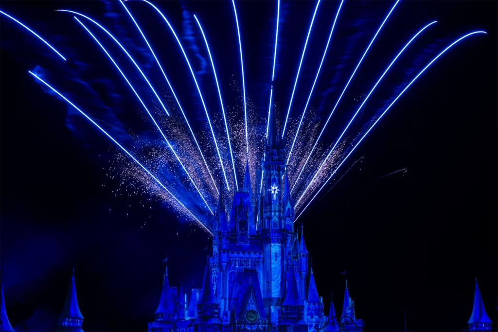 Blue and white fireworks fan out behind the illuminated castle at Magic Kingdom at night.