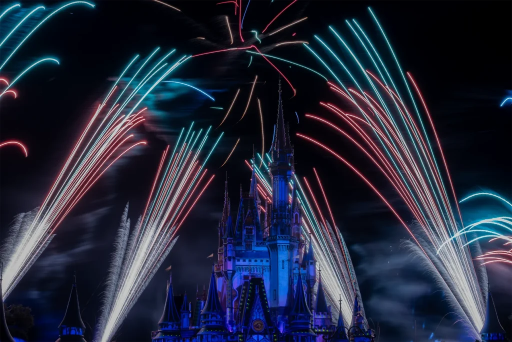 Red, cyan and white fireworks curve around the illuminated castle during a Magic Kingdom nighttime display.