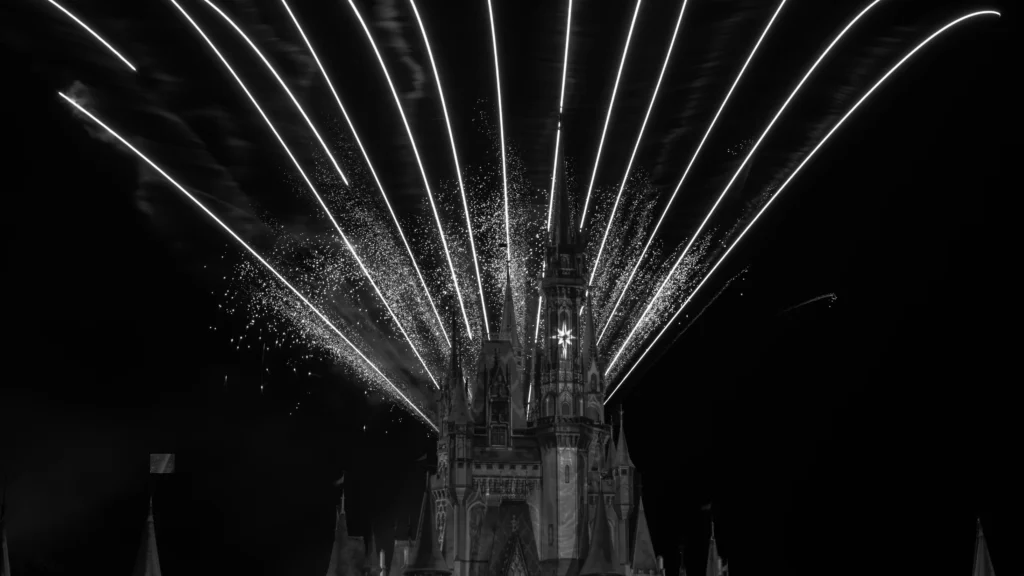 Black and white fireworks fan out behind the illuminated castle at Magic Kingdom at night.