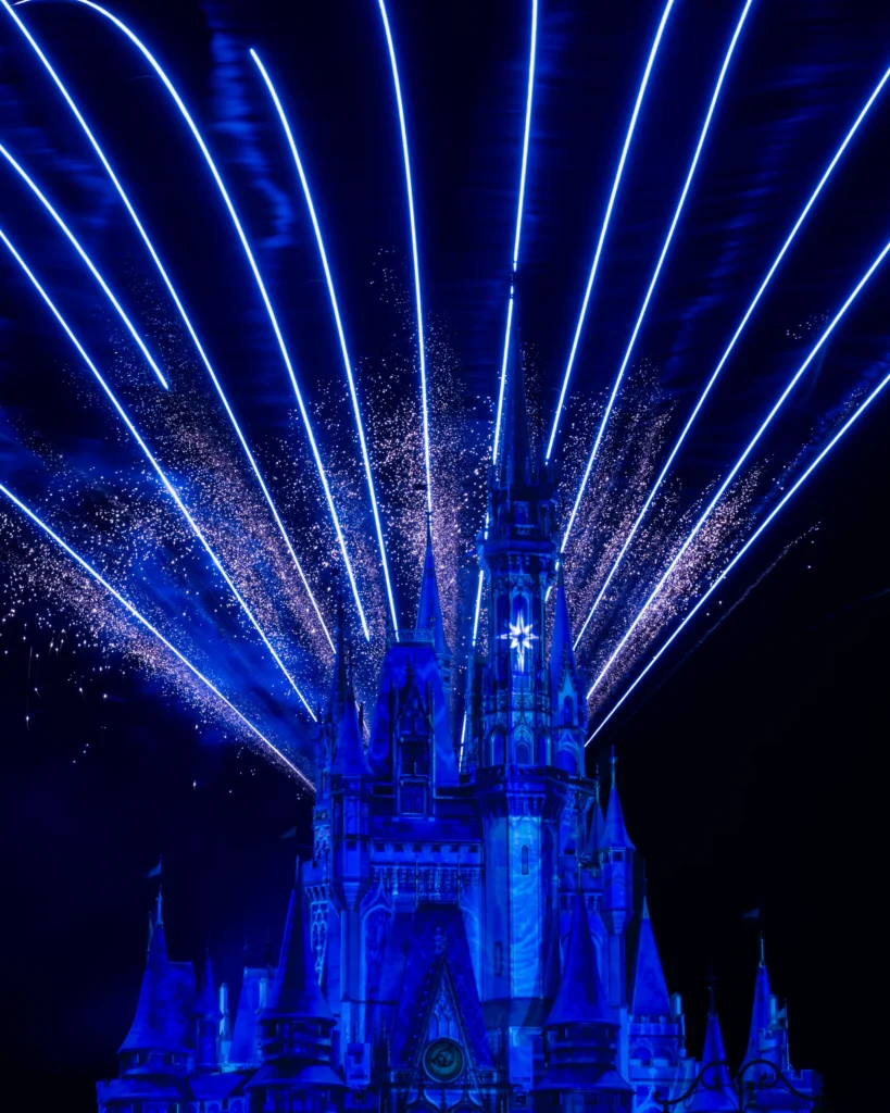 Blue and white fireworks fan out behind the illuminated castle at Magic Kingdom at night.