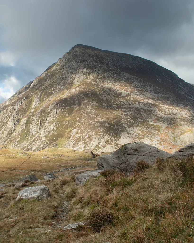 Sunlit mountain rising above a grassy valley beneath a dramatic cloudy sky.